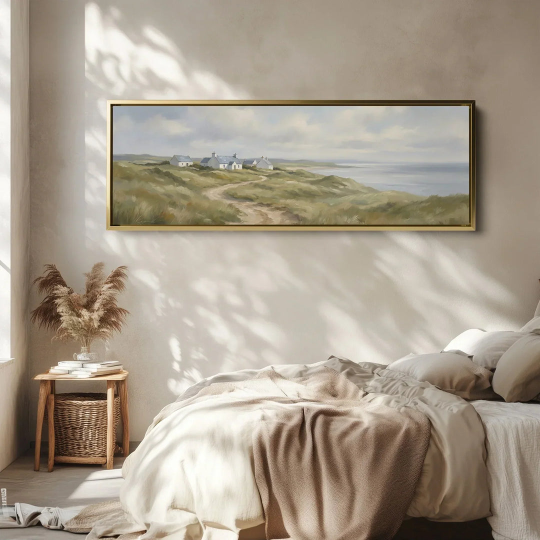 Framed long narrow horizontal coastal landscape wall art featuring seaside cottages, rolling dunes and the ocean, displayed above a bed in a modern bedroom.